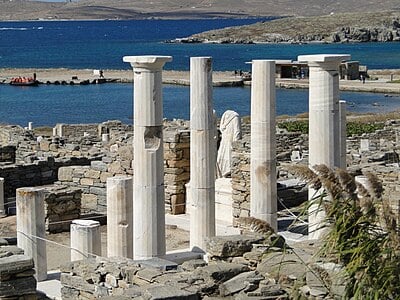 Which deity is predicted to be associated with the three conical mounds visible from Delos' Sacred Harbour?